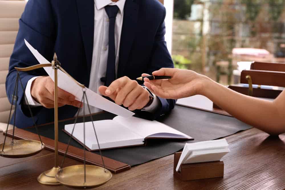 A man in a suit, holding a document and pointing at it with his pen, is seated at a desk across from another person who is handing the man a pen. On the desk are an open book, a business card holder, and a set of balance scales used by property tax experts.