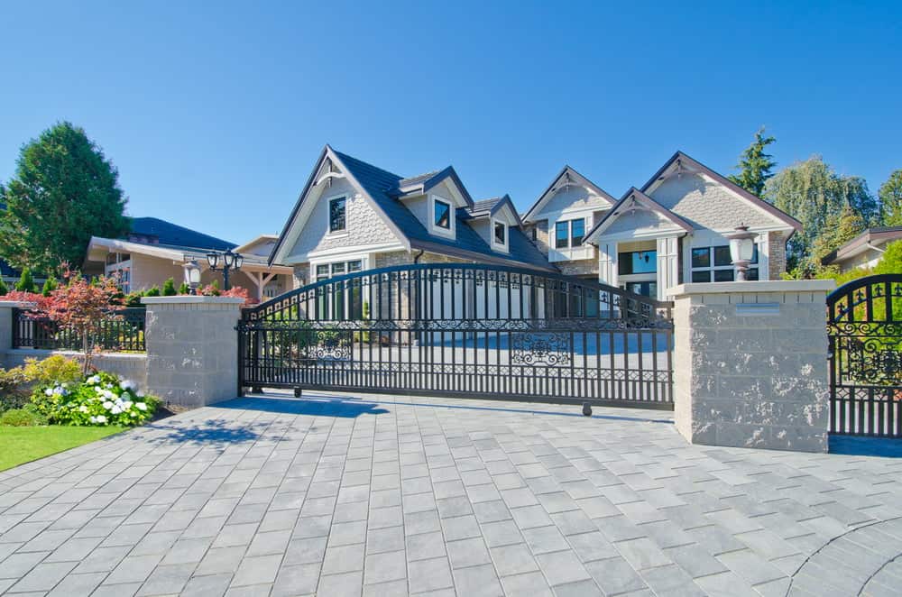 A spacious, modern home with a gray, triangular roof is surrounded by a wrought iron gate and a stone fence. The house is set against a clear blue sky, with green trees and well-manicured lawns and flower beds on either side of the driveway—an ideal haven for those seeking advice from California property tax experts.