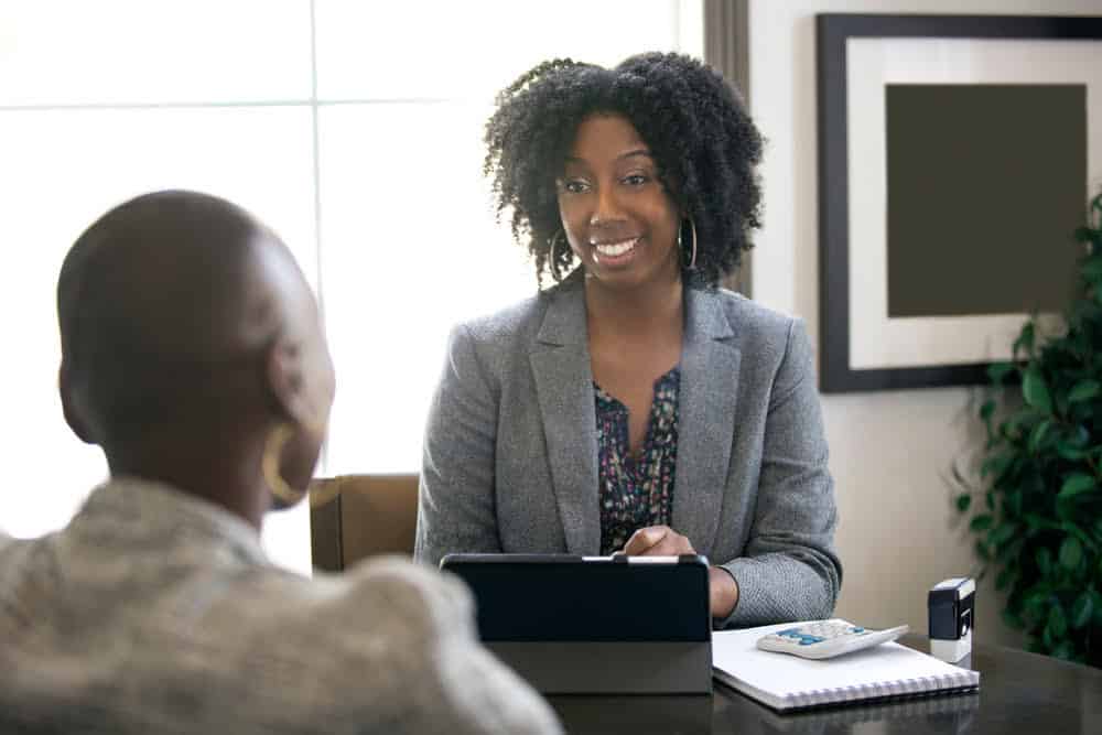 A smiling woman in a gray blazer sits at a desk with a tablet and a notebook, discussing property tax appeal strategies with another person in a light-colored shirt. The setting appears to be an office with a window and framed artwork in the background.