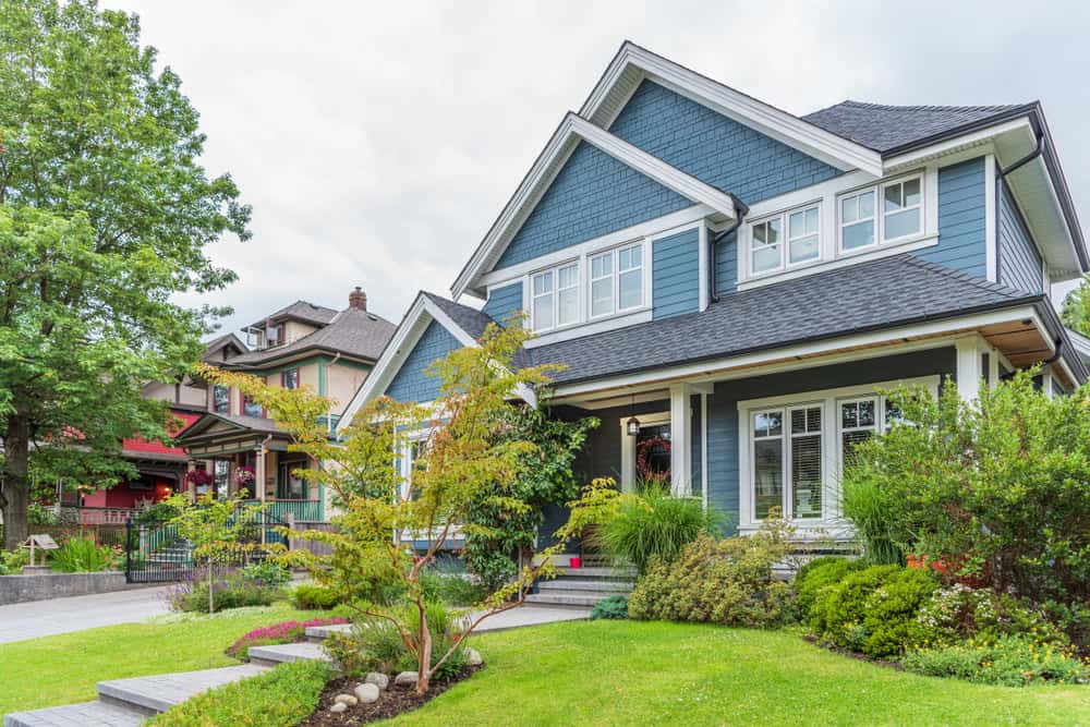 A charming two-story blue house with a gabled roof, white trim, and multiple large windows. The front yard is well-maintained with green shrubs, small trees, and a flower bed. A tree stands along the side, and neighboring houses are visible in the background—ideal for those seeking a California property tax appeal.