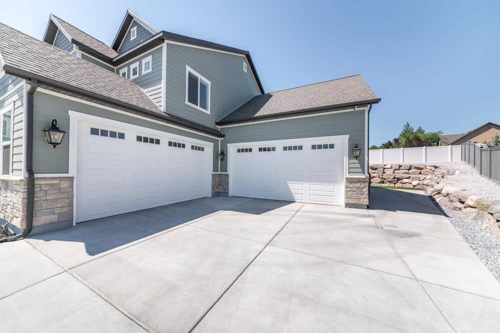 A modern home with three adjacent garage doors painted in white. The house exterior features grey siding, stone accents, and a steep roof. The wide concrete driveway leads up to the garages. A rock wall and tall wooden fence are visible on the right—a perfect property for those considering a California property tax appeal.