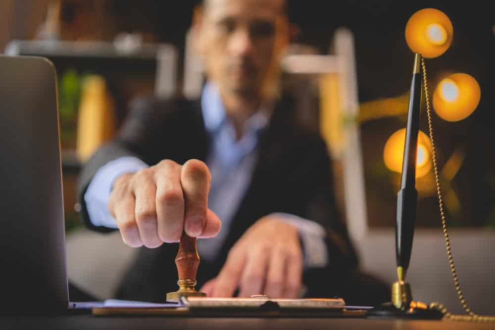 A person in a suit stamps a document on a desk, perhaps related to a California property tax appeal. The focus is on the hand and stamp, with the face blurred in the background. A laptop and a pen holder with two pens are on the desk, with warm lighting creating a professional ambiance.