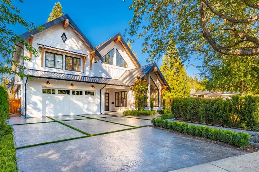 A modern two-story house with a white exterior, large windows, and a gray gable roof. The front features a spacious driveway with green lawn strips between stone slabs. Surrounded by trees and bushes against a blue sky, this home is ideal for property tax experts considering supplemental tax bills.