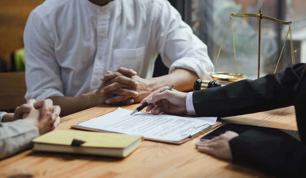 A person in a suit, likely one of the renowned property tax experts, gestures with a pen towards a document on a table as another person in a white shirt sits with folded hands. The table also holds a yellow notebook, and in the background, there is a justice scale and a gavel.