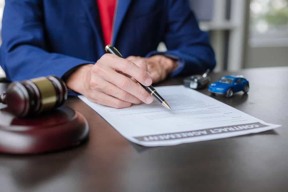 A person in a blue blazer is signing a "Contract Agreement" document on a desk. On the desk, there is a gavel and two toy cars, one silver and one blue. The background features a window with natural light, subtle reminders of property tax experts ready to assist with your Property tax appeal.