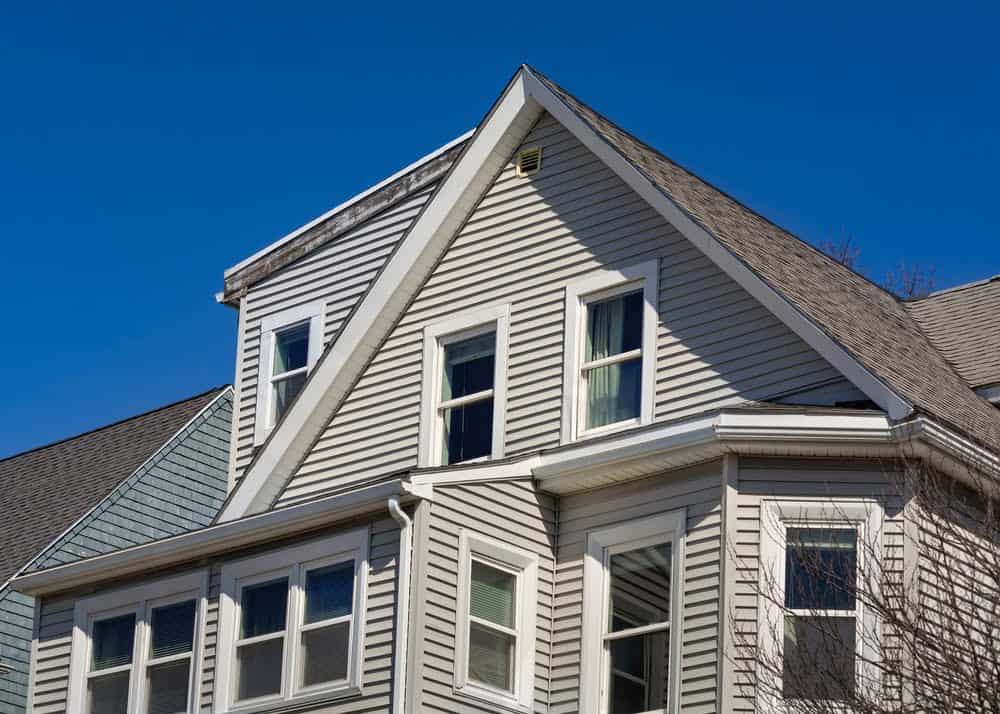 A light gray house with multiple gables and white-trimmed windows is set against a clear blue sky. The architectural design features a mix of double-hung and bay windows, highlighting a classic, multi-level structure. Trees are visible at the edge of the image, embodying the expertise of property tax experts in maximizing value.