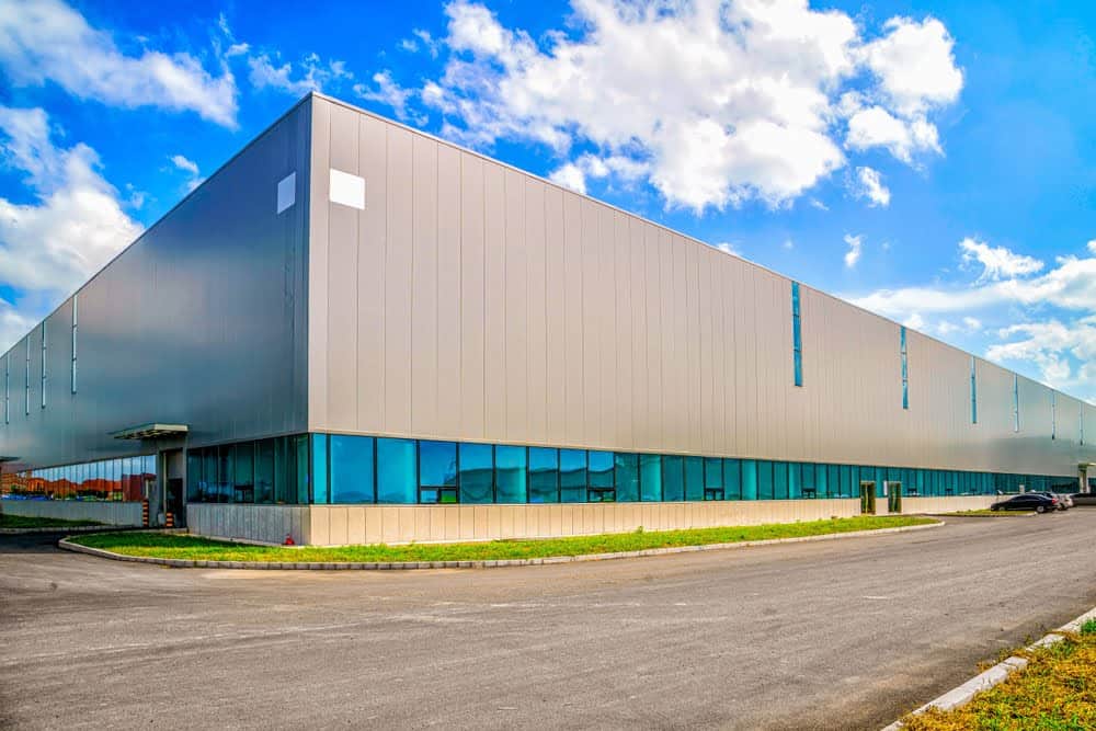 A large, modern industrial warehouse building with a sleek, metal exterior and blue-tinted windows. The property tax appeal process can be simplified by consulting property tax experts. Set against a bright blue sky with scattered white clouds, the surrounding area includes a road and some greenery.