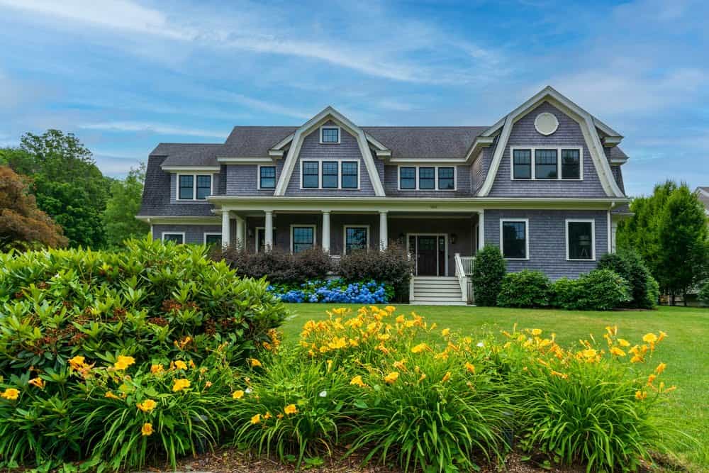 A large, two-story house with a gray exterior, white trim, and a covered porch. The home features gable roofs and several tall windows. The front yard has vibrant landscaping with yellow and blue flowers, lush green grass, and bushes under a blue sky—a true gem that property tax experts would recognize for its appeal.