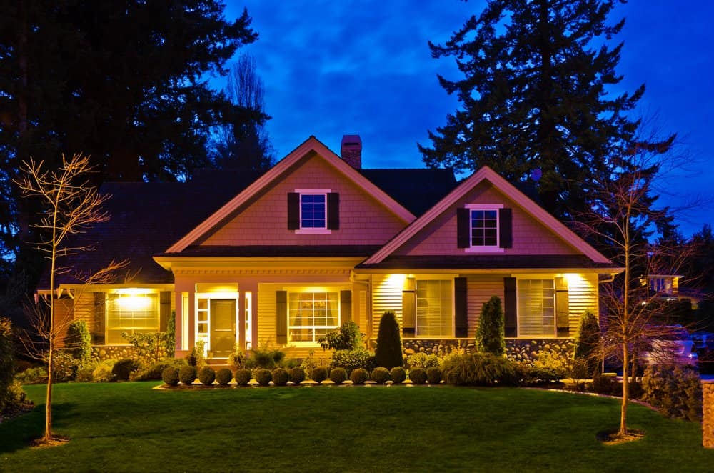 A warmly lit, two-story suburban home with a front porch, white trim, and well-maintained landscaping. The house is illuminated against a twilight sky, with trees in the background and a lush, green lawn in front. Ideal for families seeking comfort and possibly consulting property tax experts.