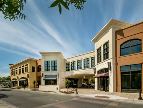 A modern commercial building complex with large windows and decorative awnings on a sunny day. The street in front is empty, and there's a clear blue sky with wispy clouds above. Property tax experts will appreciate the leafy branches from a nearby tree framing the top of the image.