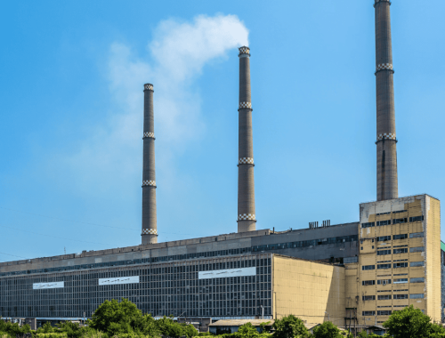 A large industrial power plant with three tall smokestacks emitting white smoke set against a clear blue sky. The expansive building has a mix of beige and gray exterior and is surrounded by a grassy area with some bushes in the foreground, reflecting its significant impact on property tax appeal considerations.