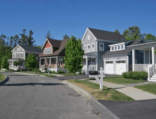 A suburban neighborhood with a street lined by several two-story houses. The houses are painted in various colors, including gray, red, and blue, and each has a well-maintained front lawn. There are trees along the street, and the sky is clear and blue—a picturesque scene ideal for any property tax expert.