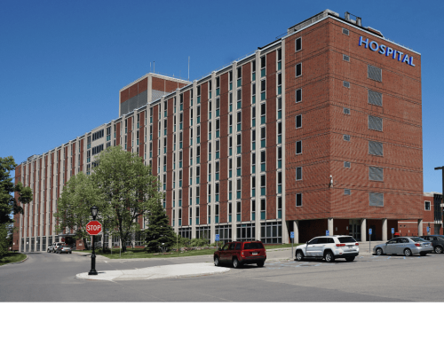 A large, multi-story red brick hospital building with a blue "HOSPITAL" sign at the top. Several parked cars are visible along the front. A stop sign and trees are present in the foreground on a sunny day. The facility also houses california property tax appeal offices.